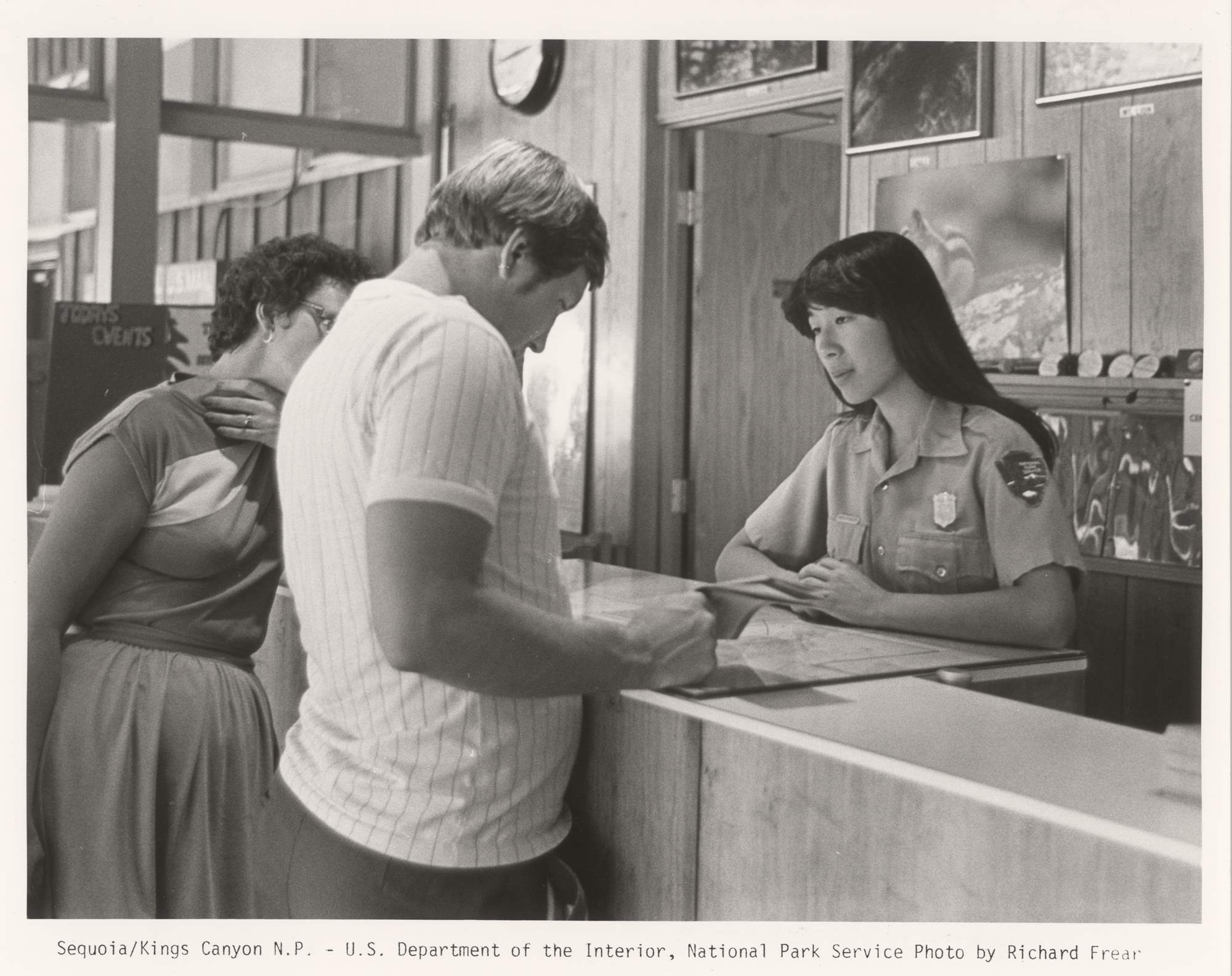 Woman in NPS uniform and shield-shaped bison badge stands behind a desk taking to visitors.